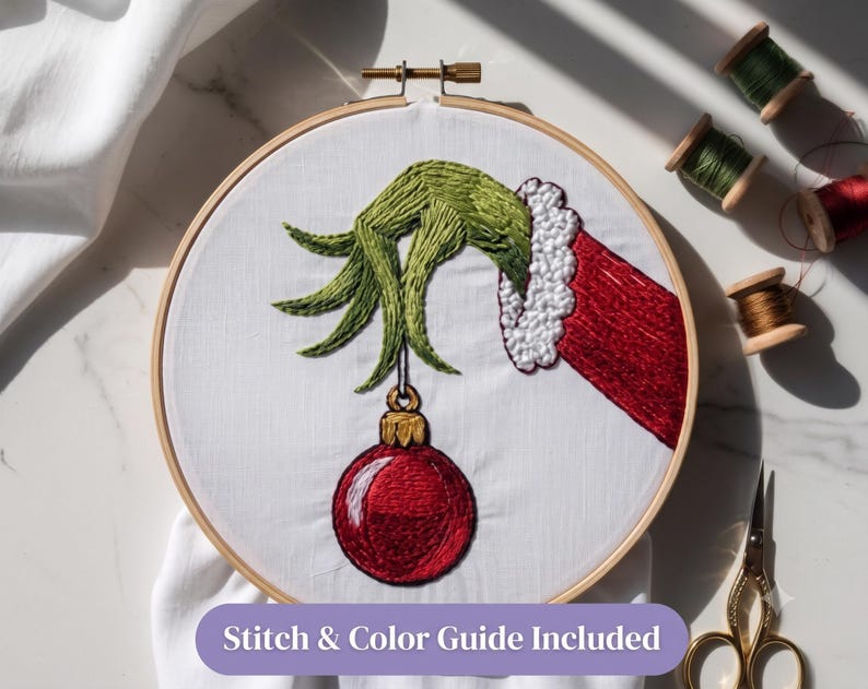 Pode incluir: Um bastidor de bordado circular de madeira contendo tecido branco com um desenho bordado &agrave; m&atilde;o de uma m&atilde;o verde de dedos longos com uma manga vermelha de borda branca, segurando um enfeite de Natal vermelho. Texto: Stitch & Color Guide Included.