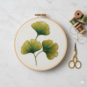 May include: An embroidery hoop with a botanical design featuring three green and yellow ginkgo leaves. Gold scissors and spools of green thread are arranged nearby, suggesting a craft project. The embroidery is on a white fabric.