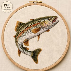 May include: A circular wooden embroidery hoop containing a detailed thread-stitched rainbow trout on off-white fabric. The fish features a green and silver body with a distinct red lateral stripe, dark spots, and yellow fins. Text reads PDF Pattern Download.