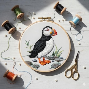 以下が含まれることがあります： 木製のフレームに入った、刺繍されたツノメドリの鳥のアート。鳥は黒、白、オレンジ色で、詳細なステッチデザインが施されています。フレームの周りには、さまざまな色の糸巻き、ビーズ、金色のハサミがあります。