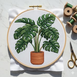 Könnte beinhalten: Stickerei einer Monstera-Pflanze in einem Terrakotta-Topf, präsentiert in einem hölzernen Stickrahmen. Die grünen Blätter und der braune Topf sind detailliert mit Faden. Grüne Garnrollen und eine goldene Schere liegen neben dem Rahmen auf einer weißen Oberfläche.