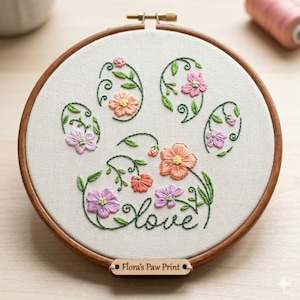May include: Embroidered paw print design with colorful flowers in a wooden hoop. The word "Love" is stitched in cursive. A small wooden tag at the bottom reads "Flora's Paw Print".