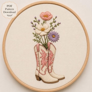 May include: An embroidery hoop containing a fabric piece with a stitched design of two cream-coloured cowboy boots with pink flame-like accents. A bouquet of pink, white, and purple flowers with green stems emerges from the boots. Text reads PDF Pattern Download.