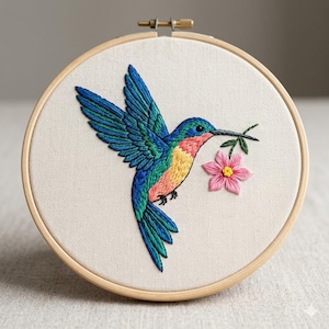 Peut inclure: Broderie d'un colibri aux couleurs vives, tenant une fleur rose dans son bec. Le colibri est brodé avec des fils bleus, verts, jaunes et roses sur un tissu crème. L'ouvrage est encadré dans un cercle en bois, pour une décoration artisanale.