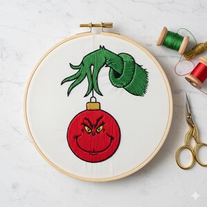May include: An embroidered hoop art piece featuring a green hand holding a red ornament with a smiling face. The embroidery is on white fabric, set in a wooden hoop. Spools of green and red thread and gold scissors are nearby.