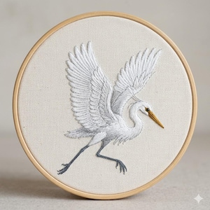 Peut inclure: Broderie d'un héron blanc en vol, encadrée dans un cercle en bois. Les ailes de l'oiseau sont déployées, révélant des détails de plumes. Le fond est en tissu neutre, et le bec de l'oiseau est doré.