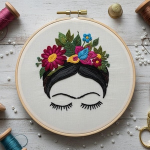 May include: An embroidered hoop art piece featuring a stylized face with closed eyes and a floral crown. The embroidery uses vibrant colors like pink, yellow, blue, and green on a cream-colored fabric. The hoop is made of wood.