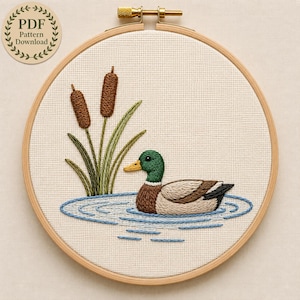 May include: A circular wooden embroidery hoop containing a cross-stitch design of a mallard duck swimming in blue water ripples. The duck has a green head, yellow beak, and brown body. Two brown cattails with green stalks are positioned nearby.