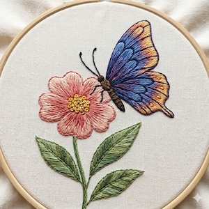 May include: Embroidered butterfly and flower design in a wooden hoop. The butterfly has blue, purple, and yellow wings. The flower is pink with a yellow center, and the leaves are green. The embroidery is on a white fabric.