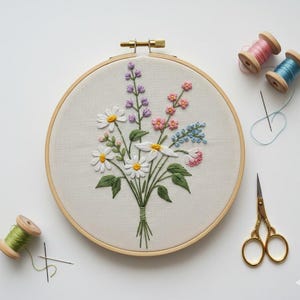 May include: A floral embroidery in a wooden hoop. The design includes white daisies, purple, pink, and blue flowers with green leaves. Pink, blue, and green thread spools, gold scissors, and needles are arranged around the hoop, ready for stitching.