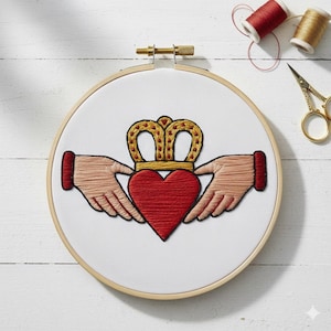 Op de afbeelding: Geborduurd Claddagh-ontwerp op witte stof in een houten borduurring. Het Claddagh-symbool toont een rood hart, een gouden kroon en twee handen. Rode en gouden garenklosjes en een gouden schaar op de achtergrond.