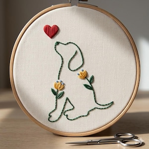 May include: Embroidered dog silhouette in a wooden hoop. The design features a green outline of a dog, a red heart, and yellow flowers with green leaves. A pair of small scissors rests below the hoop.