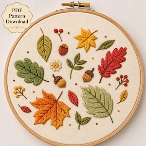 Puede incluir: Un bastidor de bordado con tela color crema que presenta un patrón bordado de hojas de otoño, bellotas y bayas en tonos rojos, naranjas, amarillos y verdes. Una pequeña insignia en la esquina superior izquierda muestra el texto PDF Pattern Download.