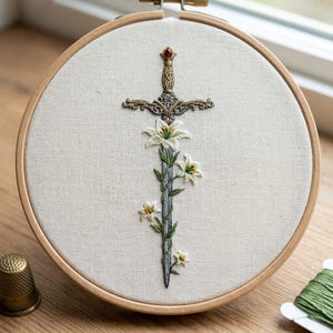 May include: Embroidered artwork in a wooden hoop. The design features a sword with a floral stem and white flowers. The sword has a decorative hilt with a red gem. The background is a light beige fabric.