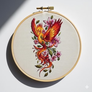 Puede incluir: Un bordado detallado de un fénix con plumas vibrantes en naranja, rojo y amarillo, rodeado de flores rosas y hojas verdes. La obra de arte está en un bastidor de bordado redondo de madera, creando una pieza decorativa.
