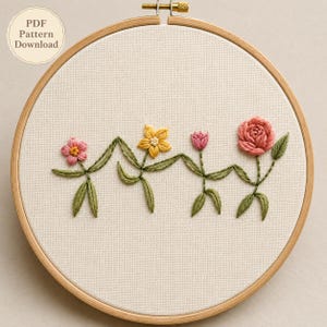 Può includere: Un telaio da ricamo circolare in legno contenente tessuto bianco con un disegno floreale cucito a mano. Il ricamo presenta quattro fiori distinti in tonalità rosa, giallo e rosso, collegati da steli e foglie verdi. Il testo recita PDF Pattern Download.