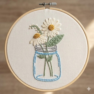May include: A circular wooden embroidery hoop containing off-white fabric with a hand-stitched design of two white daisies with yellow centers and green leaves inside a light blue glass mason jar, featuring a metal tightening screw at the top.