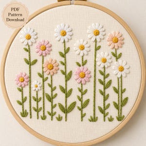 Può includere: Un telaio da ricamo contenente un pannello in tessuto con fiori cuciti a mano in bianco, rosa chiaro e giallo. Il design presenta vari fiori simili a margherite su steli verdi con foglie. Un badge circolare riporta PDF Pattern Download.