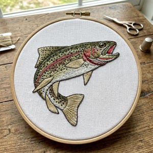 May include: Embroidered rainbow trout on white fabric stretched within a wooden embroidery hoop. The fish features detailed stitching in shades of green, gold, red, and brown. A pair of small scissors and a thimble are visible nearby.