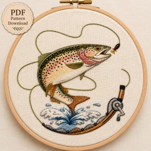 May include: An embroidery hoop containing a fabric panel with a stitched design of a rainbow trout leaping from water. The fish features green, pink, and cream thread details. A fishing rod handle and reel are stitched below with a line and lure.
