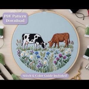Op de afbeelding: Een borduurring met lichtblauwe stof en een handgeborduurde scène van een zwart-witte koe en een bruine koe die grazen in een veld met kleurrijke wilde bloemen. De tekst PDF Pattern Download en Stitch & Color Guide Included is zichtbaar.
