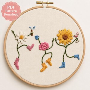 Könnte beinhalten: Ein runder Stickrahmen aus Holz mit einem weißen Stoffeinsatz, auf dem handgestickte Blumen mit Stiefeln zu sehen sind. Eine Gänseblume, eine rosa Blume und eine Sonnenblume tanzen und halten eine Flasche, einen blauen Becher und ein Cocktailglas.