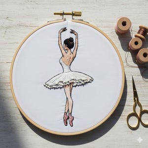 Puede incluir: Obra de arte bordada de una bailarina con tutú blanco y zapatillas de punta rosas, con los brazos levantados. El diseño está en un bastidor de bordado de madera. Se ven carretes de hilo adicionales y tijeras doradas.