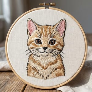 May include: An embroidery hoop containing a fabric panel with a detailed thread-stitched portrait of an orange tabby cat. The cat features brown eyes, pink-tinted ears, and black whiskers, centered on a plain off-white fabric background.