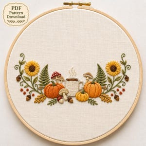 以下が含まれることがあります： 刺繍枠にはめ込まれた布地に、2つのオレンジ色のカボチャ、湯気の立つ白いマグカップ、黄色のヒマワリ、緑色のシダ、茶色のドングリ、様々なキノコが刺繍されたデザイン。クリーム色の背景。PDF Pattern Download。