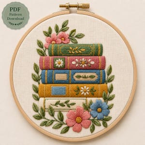 May include: A circular wooden embroidery hoop containing a fabric panel with a hand-stitched design of a stack of five books in green, pink, blue, yellow, and cream, surrounded by embroidered green leaves and pink and blue flowers.