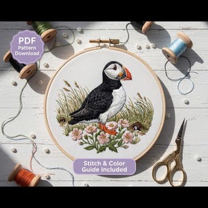 以下が含まれることがあります： 刺繍枠には、パフィン、緑の草、ピンクの花が刺繍された白い布地が入っています。テキストには「PDF Pattern Download」および「Stitch & Color Guide Included」と書かれています。糸巻きと金属製のハサミが含まれています。