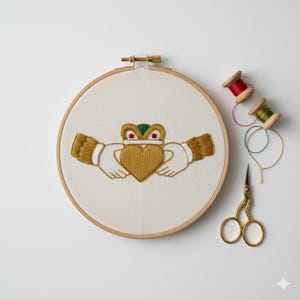 Op de afbeelding: Geborduurd Claddagh-ontwerp in een houten hoepel. Het Claddagh-symbool toont handen die een hart vasthouden, bekroond met een kroon, in gouddraad. Extra garenklosjes en een gouden schaar liggen naast de hoepel.