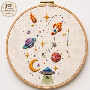 Peut inclure: Un tambour à broder contenant un tissu crème avec un motif sur le thème de l'espace comprenant une fusée, une planète avec des anneaux, un croissant de lune, un OVNI, un astronaute, des étoiles et une comète. Le texte PDF Pattern Download est présent.