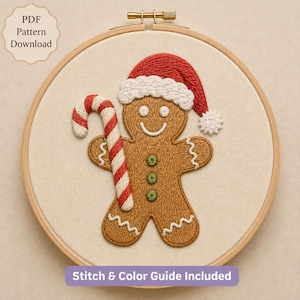May include: A circular wooden embroidery hoop containing a fabric panel with a stitched gingerbread person wearing a red and white Santa hat. The figure holds a red and white candy cane. Text reads: PDF Pattern Download and Stitch & Color Guide Included.