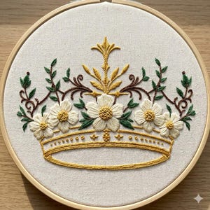 May include: Embroidered crown design in a wooden hoop. The crown is yellow with white flowers, green leaves, and brown vines. The embroidery is on a cream-colored fabric.