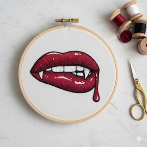 May include: Embroidery hoop featuring a detailed, red-lipped vampire mouth with fangs and a drop of blood. The artwork is set against a white background within a wooden hoop. Spools of thread and gold scissors are visible.