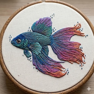 Puede incluir: Un pez Betta bordado en un aro de madera. El pez presenta puntadas detalladas en tonos azules, turquesas, morados y naranjas. Pequeñas burbujas con cuentas rodean al pez sobre una tela color crema.