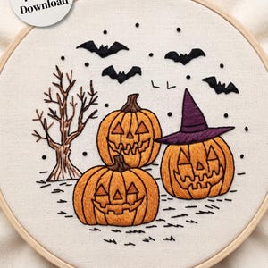 Puede incluir: Un bastidor de bordado que contiene una pieza de tela con una escena de Halloween cosida a mano, con tres calabazas naranjas, una con un sombrero de bruja morado, un árbol marrón sin hojas y murciélagos negros sobre un fondo blanco con texto.