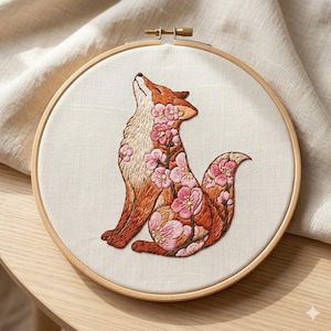 May include: Embroidered fox hoop art. The fox is depicted in shades of orange and brown, with pink floral accents. The fox is looking up, and the embroidery is set in a wooden hoop. The background is a light beige fabric.