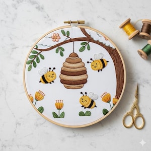May include: Embroidered hoop art with a bee theme. The design shows a brown beehive, three smiling bees, yellow flowers, and green leaves on white fabric. The embroidery is framed in a wooden hoop, perfect for home decor.