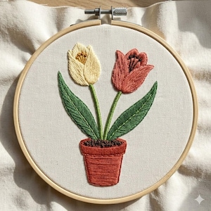 May include: Embroidered floral hoop art featuring two tulips in a terracotta pot. One tulip is cream-colored, the other is red. Green leaves and stems are visible. The embroidery is set in a wooden hoop.