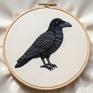 May include: A circular wooden embroidery hoop containing a piece of off-white fabric. Centered on the fabric is a detailed black thread embroidery of a crow standing in profile, facing right, with textured feather details and a sharp beak.