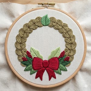 May include: A circular embroidery hoop containing a piece of off-white fabric with a hand-stitched wreath design. The wreath is formed by braided tan thread, accented at the bottom with a bright red bow, green leaves, and small red berries.