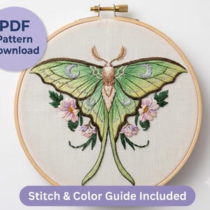 Op de afbeelding: Een borduurring met een stuk stof met een handgeborduurde maannachtvlinder in groentinten, voorzien van zilveren maansikkelaccenten en roze bloemen. Een paarse cirkel toont PDF Pattern Download en een paarse balk Stitch & Color Guide Included.