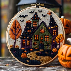 Op de afbeelding: Een ronde borduurring met een geborduurde scène van een spookhuis met oranje ramen, een kronkelend pad en kleine pompoenen. Het ontwerp bevat zwarte vleermuizen in de lucht, twee oranje bomen en een paars met groen huis als centraal element.
