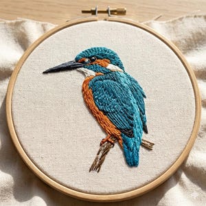May include: A circular wooden embroidery hoop containing a piece of off-white fabric with a detailed thread-stitched kingfisher bird. The bird features vibrant blue, orange, and white plumage, a long dark beak, and is perched on a brown branch.
