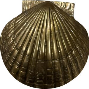May include: A brass-coloured, seashell-shaped decorative object. The shell has a ridged, fan-like design with a rectangular top. The surface has a shiny, metallic finish. Likely a decorative piece for a coastal or nautical theme.