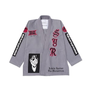 Pode incluir: Um gi cinzento de artes marciais com detalhes bordados em vermelho e preto. O gi apresenta o texto "SJR" em vermelho na frente e "Rebels Against The Mainstream" na parte inferior. Patches e logotipos são visíveis nas mangas e no peito.