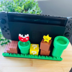 以下が含まれることがあります： 黒いNintendo Switchコンソールが装飾スタンドにドッキングされています。スタンドには、緑色のパイプ、クエスチョンマークブロック、赤いキノコなど、スーパーマリオビデオゲームシリーズのカラフルな3Dプリント要素が使用されています。