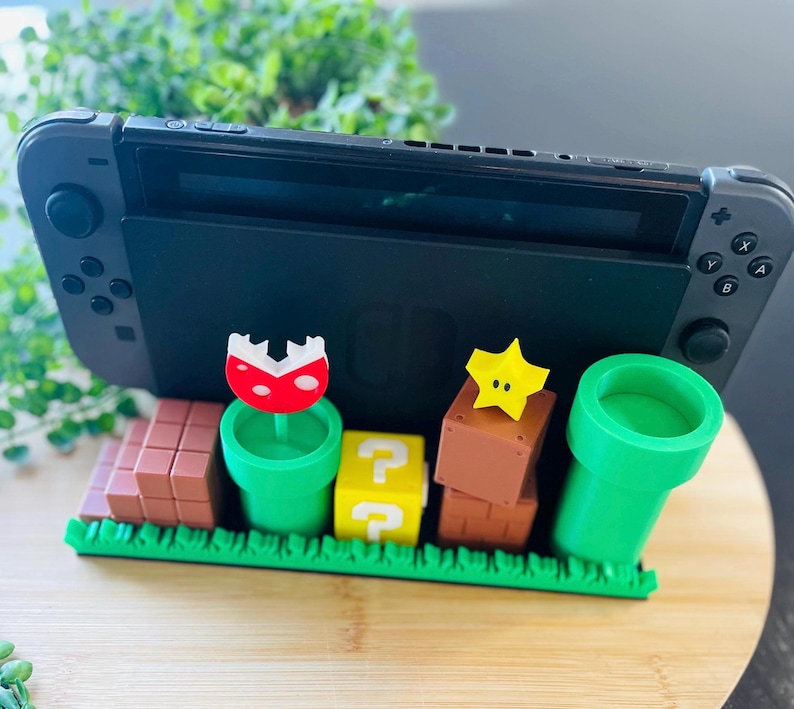 May include: A black Nintendo Switch console is docked on a decorative stand. The stand features colorful 3D-printed elements from the Super Mario video game series, including a green pipe, question mark block, and a red mushroom.
